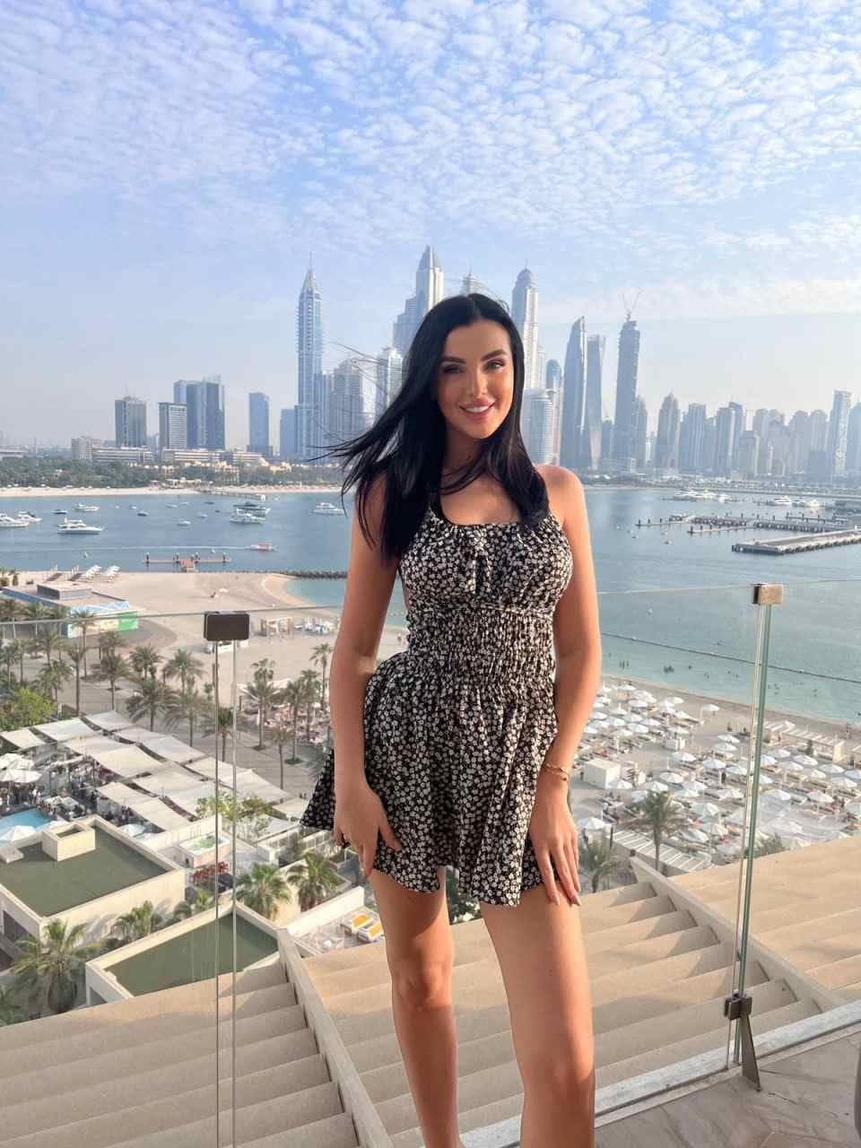 Standing on an outdoor staircase, she wears a black-and-white floral dress, with a city skyline and beach in the background.