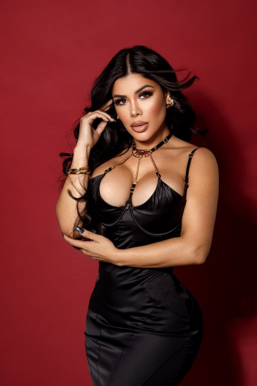 Woman in black satin slip dress with plunging neckline and gold jewelry, standing against red backdrop, one hand in hair, other on chest.