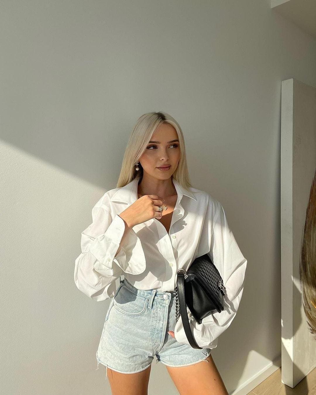Against a plain wall, she wears an oversized white button-up shirt and denim cutoffs, holding a black crossbody bag while looking aside.