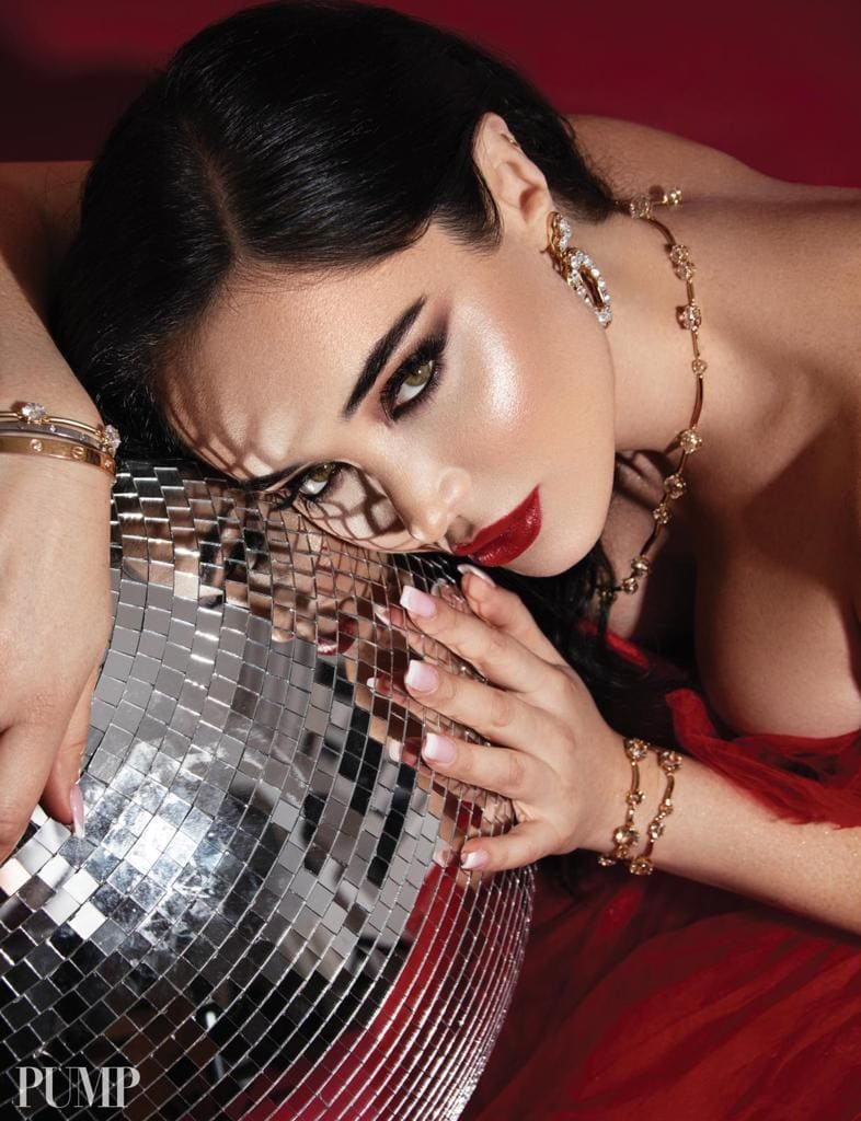 Woman in a red strapless gown and gold jewelry resting her head on a mirrored disco ball against a red background.